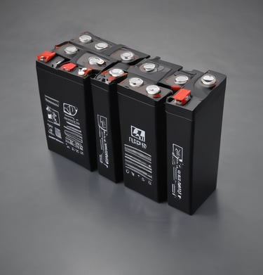 A lithium battery labeled '12V 100Ah' rests on a concrete bench in an outdoor setting with trees and sunlight visible. Wires are attached to the battery, and beside it is a white device, possibly a charger.