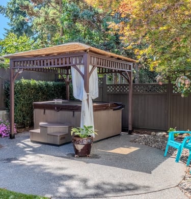 A backyard hot tub under a wooden gazebo with blue Adirondack chairs on a stone patio.