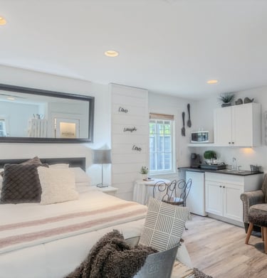 Modern studio apartment featuring a white bed, large wall mirror, and compact kitchenette with white cabinets.