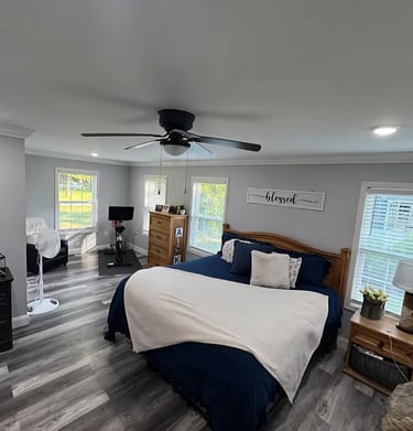 Modern master bedroom with grey LVP flooring, large windows for natural light, and a ceiling fan. 