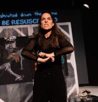A white woman signs emphatically "you will not be resuscitated" in front of a painting and text
