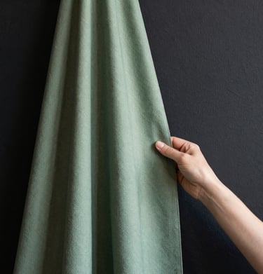 Minimalist close-up of a designer's hand holding a sage green textile swatch against a charcoal black wall, soft natural light.