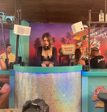 Kat Bendis performing at a vibrant Ibiza beach club bar with tropical decor and neon signs.