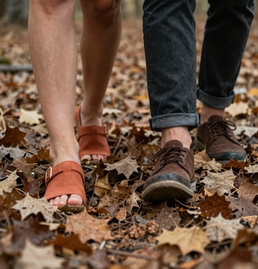 A close-up shot of a couple's feet walking through dry leaves in a North American forest. Earthy terracotta, charcoal, and soft sand colors in a cinematic style.