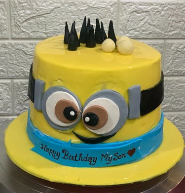 Yellow Minion birthday cake for a son with goggles and a blue ribbon banner on a cake stand.
