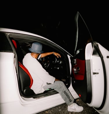 A person in streetwear sitting in a white luxury car with red leather interior at night.