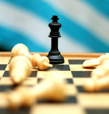 Black king chess piece standing victorious among fallen white pieces on a wooden game board.
