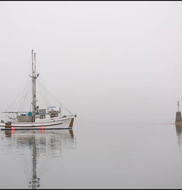 A white commercial fishing boat floats on calm, foggy water near a stone lighthouse beacon.