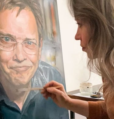 Stefanie placing final touches on oil portrait painting of smiling man with glasses.