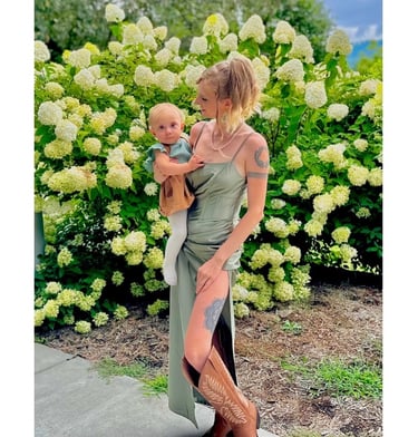 Jade in a green dress and boots holding baby Nova by flow bush, representing maternal instinct and caregiving