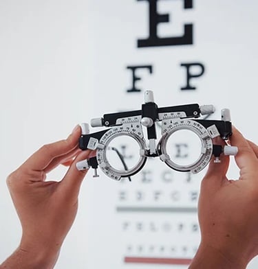 Mãos segurando um foróptero em frente a uma tabela de acuidade visual durante exame oftalmológico