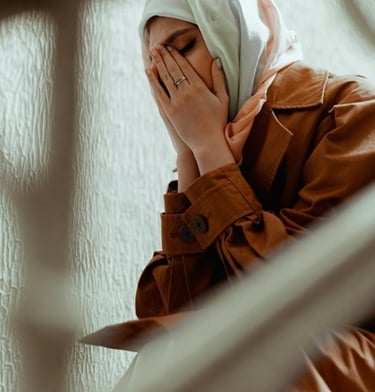 A woman in a hijab and brown coat sits on stairs covering her face in distress or prayer.
