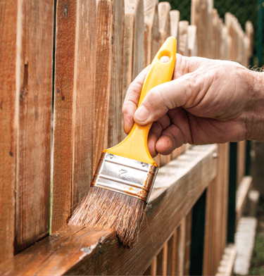 Close-up of a landscaper applying protective wood sealer on a cedar fence post with a brush.