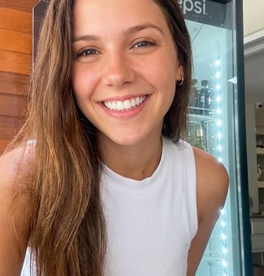 A smiling young woman with long brown hair wearing a white ribbed crop top standing in front of a cooler.
