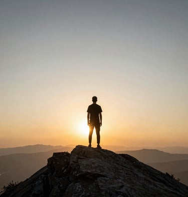 A single figure standing on a mountain peak looking at a vast, warm sunrise, expansive sky.