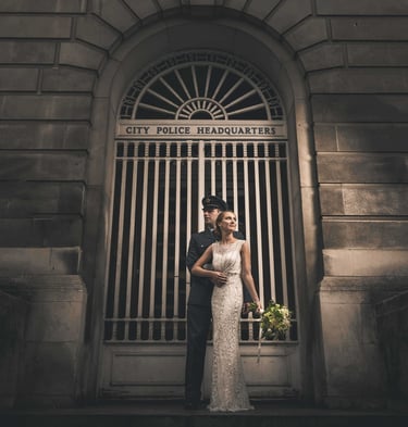 A bride in a white gown and a groom in a police uniform pose outside City Police Headquarters.