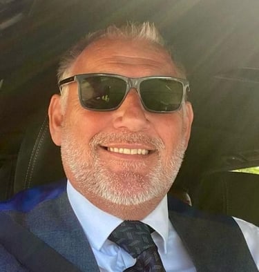 Nigel, smiling with a grey beard wearing sunglasses, a grey waistcoat, and a patterned tie.