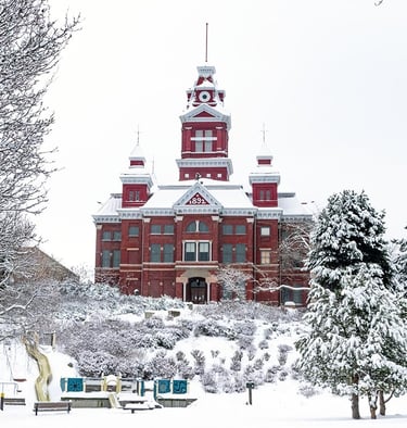 Historic red brick clock tower building standing behind a snow-covered winter park and playground.