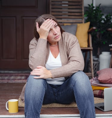 Middle-aged woman sitting on her front porch, hand on forehead, looking tired and reflective. A lapt
