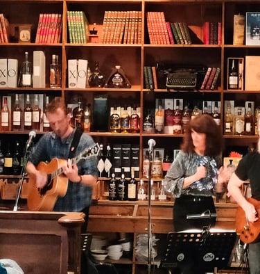 Live folk band with acoustic guitars performing in a rustic pub with shelves of liquor bottles.