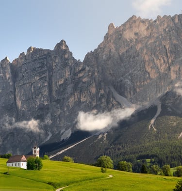 A small white chapel sits on a lush green meadow beneath the jagged peaks of the Italian Dolomites.