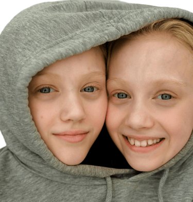 Two smiling girls with blue eyes sharing a single grey hoodie
