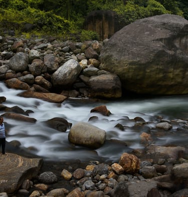 Picture of a river water caught in long exposure surrounded by big boulders on which there is a man.