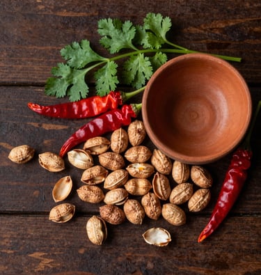 A rustic, top-down photograph of a dark wooden surface. Artistically arranged on it are high-quality, whole ingredients from tribal farmers: a scattering of raw groundnuts in their shells, vibrant red chilis, and fresh green coriander leaves next to a small terracotta bowl. The lighting is warm and natural, emphasizing organic textures and earthy colors of brown, forest green, and rust orange.
