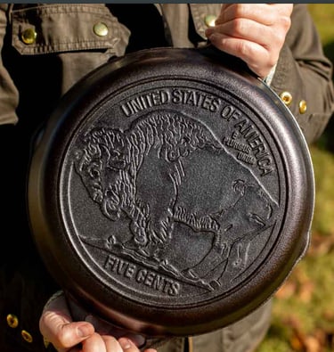 Cast iron skillet featuring a detailed Buffalo Nickel design on the bottom being held outdoors.