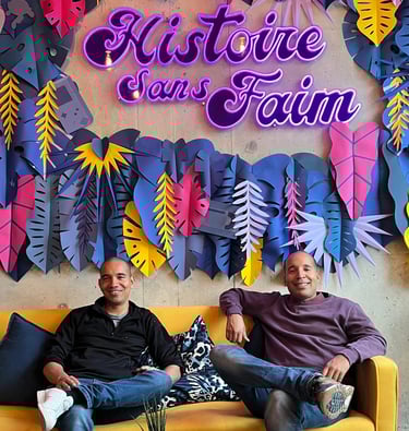 Christopher et Michael assis dans un canapé jaune sous le néon violet Histoire Sans Faim, décor pop coloré.