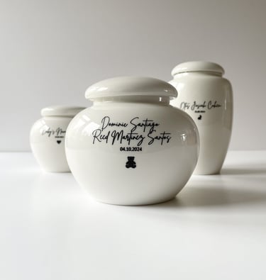 Set of three glossy white ceramic baby urns, each engraved with names, dates, and tiny black heart or bear icons.
