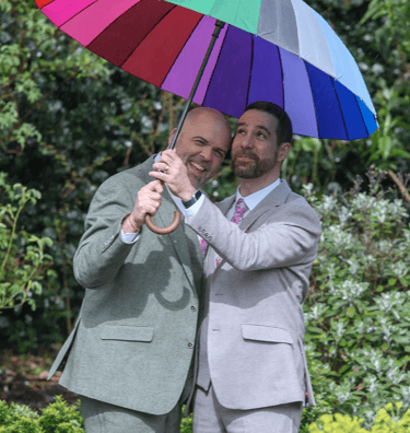 two grooms standing next to each other holding a rainbow colored umbrella