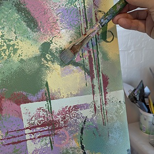 hand holding paintbrush against painting in greens, pinks, yellows, purples