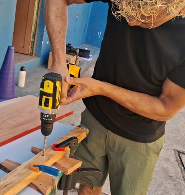 A woodworker uses a cordless electric power drill to bore holes in a wooden plank secured with clamps.