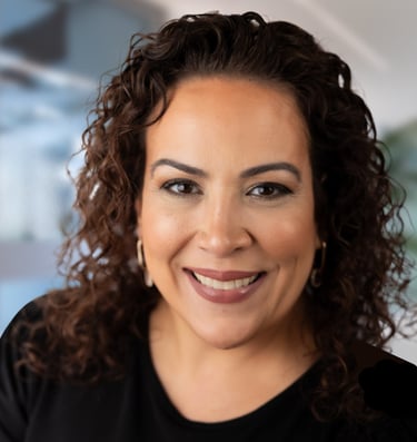 Professional headshot of a smiling woman with curly brown hair wearing a black shirt and hoop earrings.