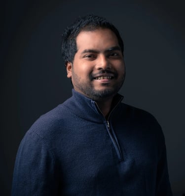 Professional headshot of Tharinda H.A smiling in a navy sweater against a dark gray background.