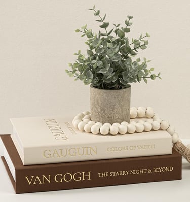 Potted artificial eucalyptus plant on a stack of decorative Van Gogh and Gauguin art books with wooden beads.