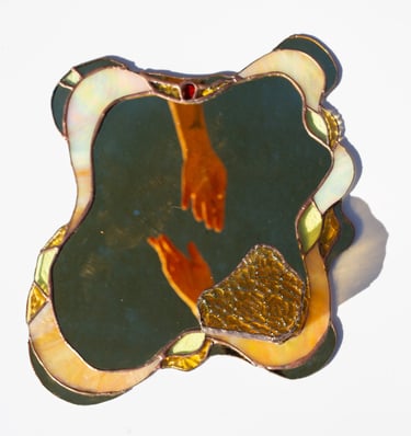 Espejo artesanal de vitral ondulado con borde iridiscente y el reflejo de unas manos extendiéndose