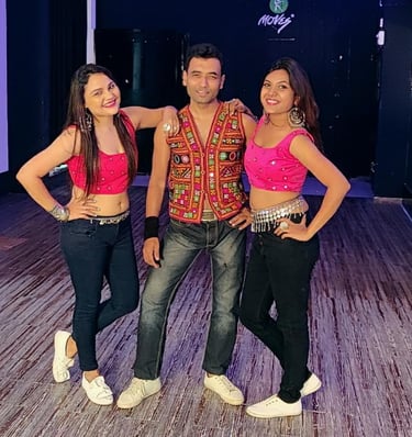 Three Bollywood dancers pose in a studio wearing colorful traditional embroidered vests and pink crop tops.