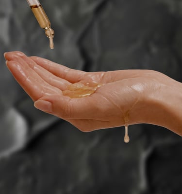 Golden facial oil dripping from a glass dropper into a person's open palm against a dark stone background.
