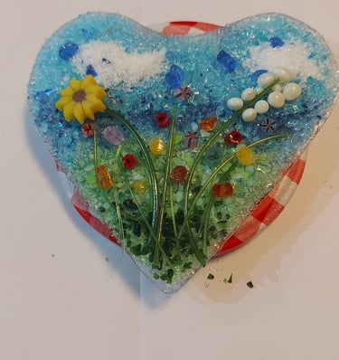 Handmade heart-shaped fused glass art featuring a floral meadow with a yellow sunflower and blue sky.