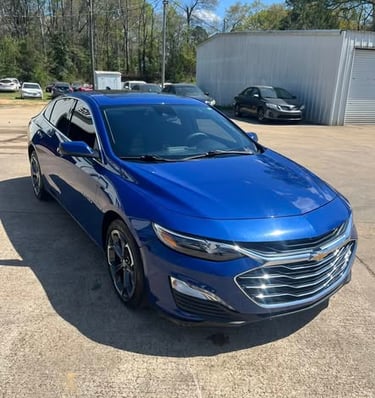 A bright blue Chevrolet Malibu sedan parked on a sunny concrete lot at a car dealership.