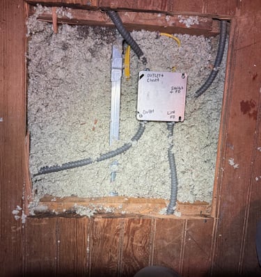 Electrical junction box with conduit wiring installed in blown-in attic insulation under wooden floorboards.