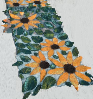 Handmade nuno felted scarf featuring yellow sunflowers and green leaves on a white wooden surface.