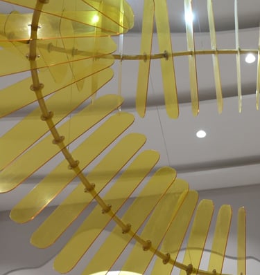 Yellow acrylic kinetic sculpture hanging from a white ceiling in a modern architectural interior.