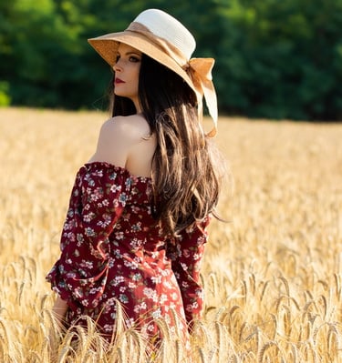 a beautiful woman in a dress and hat standing in a wheat field summertime
