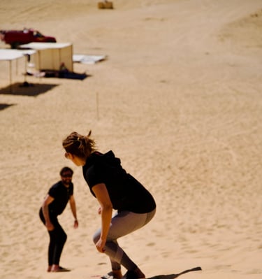 A person sandboarding run down a sand dune. Another person, standing in the background.