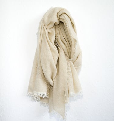 Soft cream-colored pashmina schal with fringed edges, elegantly draped on a minimal white wall.