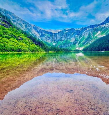 Clear alpine lake reflecting rugged mountains and green forests under a blue sky.