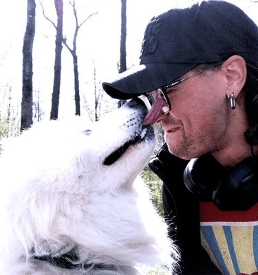 a photo of a man wearing a black baseball cap and glasses being licked in the face by white dog